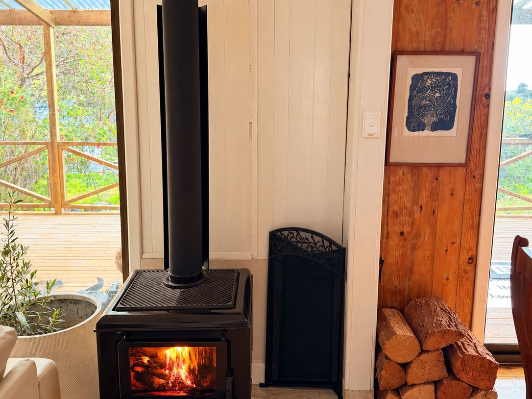 Wood heater lit with firewood stacked beside it at Sandy Feet Retreat